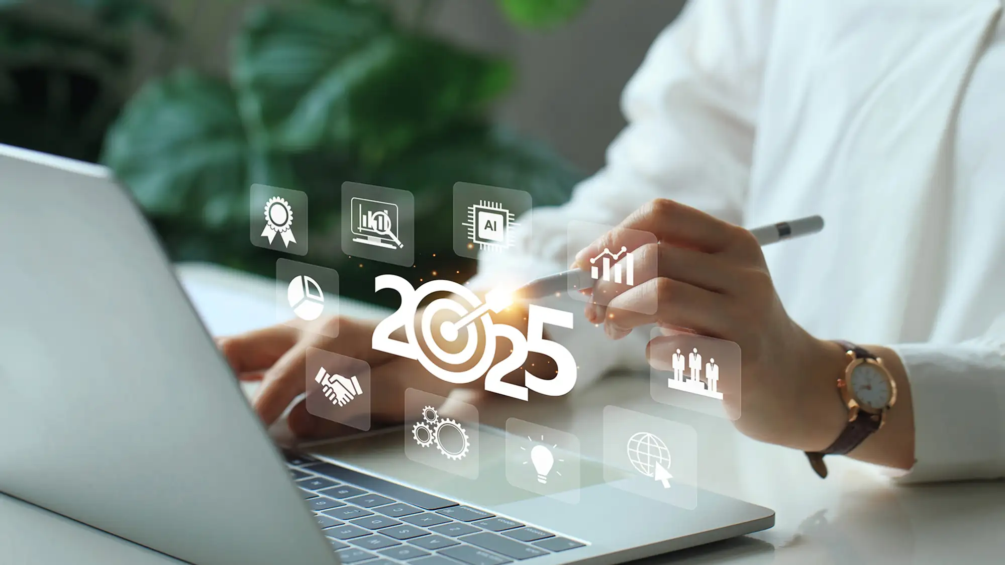 A person sitting at a laptop using a stylus to create a stylized 2025 surrounded by various business-related icons